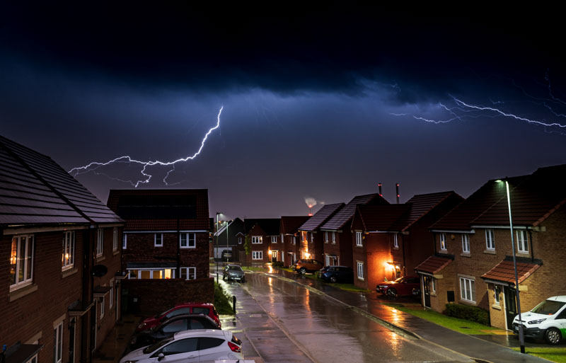 A vivid lightning strike illuminating an urban landscape at night, featuring houses and cars